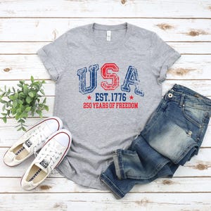 May include: A gray t-shirt with the word "USA" in red and blue distressed lettering, with "EST. 1776" and "250 YEARS OF FREEDOM" printed below. The shirt is paired with blue jeans and white sneakers.