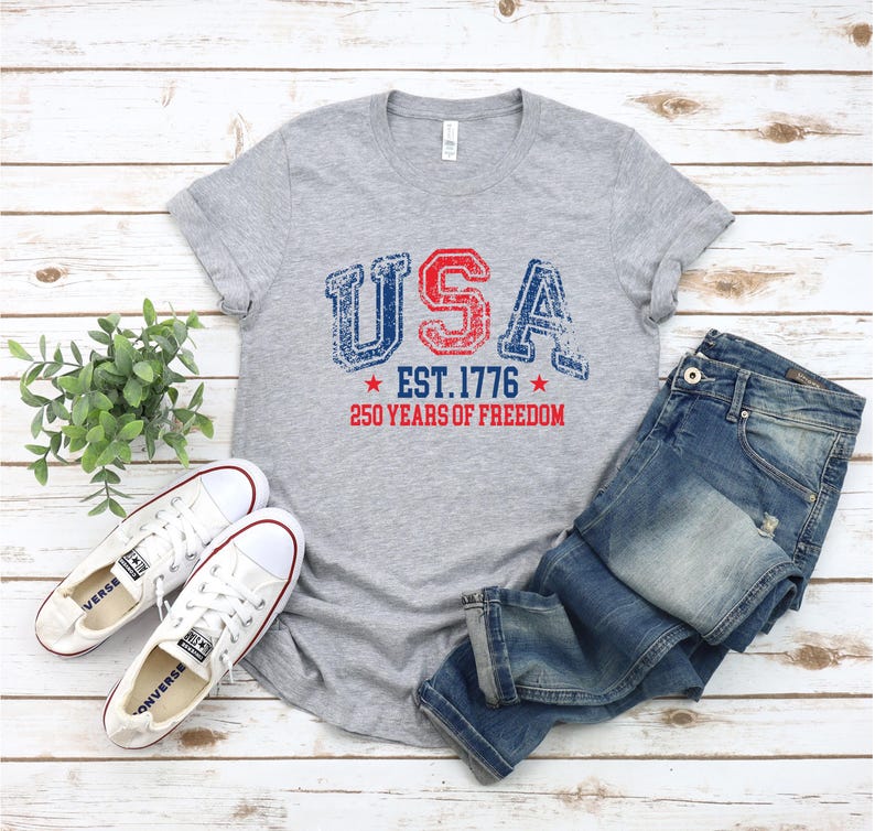 May include: A gray t-shirt with the word "USA" in red and blue distressed lettering, with "EST. 1776" and "250 YEARS OF FREEDOM" printed below. The shirt is paired with blue jeans and white sneakers.