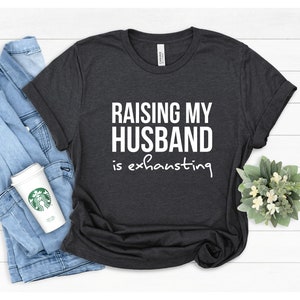 May include: A dark gray t-shirt with the text "Raising my husband is exhausting" printed in white.