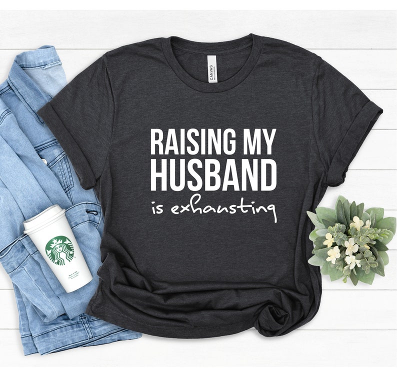 May include: A dark gray t-shirt with the text "Raising my husband is exhausting" printed in white.