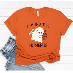 May include: Orange t-shirt with a white ghost holding a bone and a spider web. The text on the shirt reads "I found this humerus".
