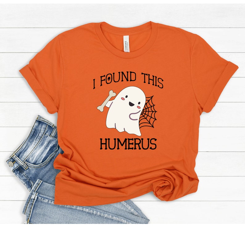 May include: Orange t-shirt with a white ghost holding a bone and a spider web. The text on the shirt reads "I found this humerus".