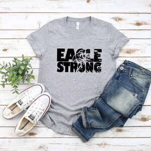 Op de afbeelding: Grijs T-shirt met zwarte tekst die "Eagle Strong" zegt en een afbeelding van een adelaar in een basketbal.