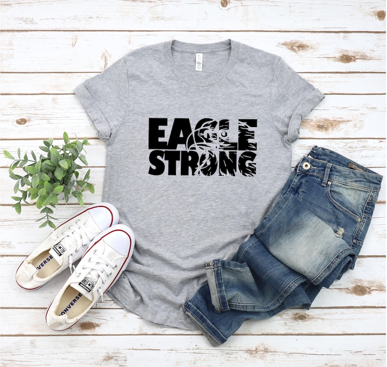 Op de afbeelding: Grijs T-shirt met zwarte tekst die "Eagle Strong" zegt en een afbeelding van een adelaar in een basketbal.