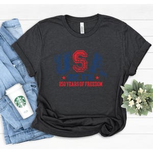 May include: Dark gray t-shirt with the text "USA" in blue and red, along with "250 YEARS OF FREEDOM". The shirt is folded, with a denim jacket and a coffee cup visible in the background. A small floral arrangement is also present.