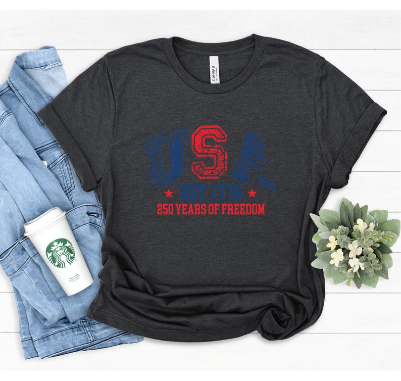 May include: Dark gray t-shirt with the text "USA" in blue and red, along with "250 YEARS OF FREEDOM". The shirt is folded, with a denim jacket and a coffee cup visible in the background. A small floral arrangement is also present.