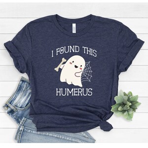May include: A navy blue t-shirt with a white ghost holding a bone and a spider web. The text "I found this humerus" is printed on the shirt.