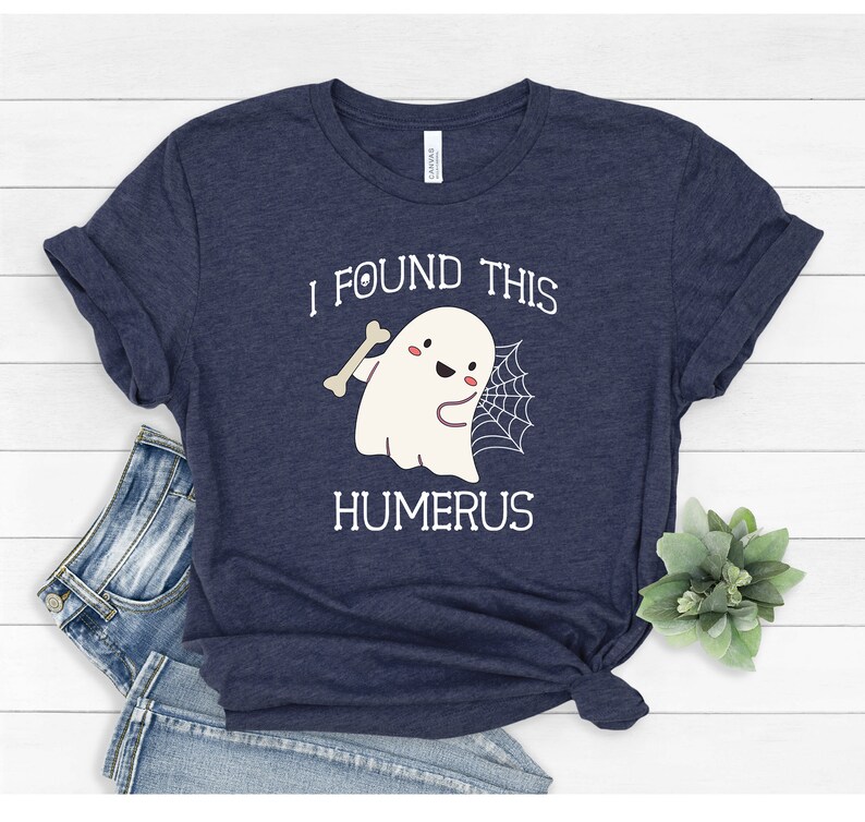 May include: A navy blue t-shirt with a white ghost holding a bone and a spider web. The text "I found this humerus" is printed on the shirt.