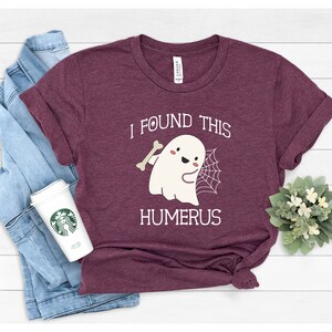May include: A maroon t-shirt with a white graphic of a ghost holding a bone and a spider web. The text "I found this humerus" is printed below the ghost.