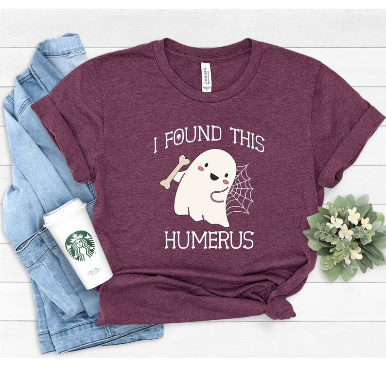 May include: A maroon t-shirt with a white graphic of a ghost holding a bone and a spider web. The text "I found this humerus" is printed below the ghost.