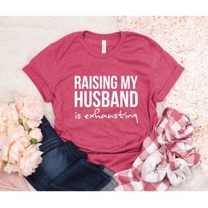 May include: A red t-shirt with the text "Raising my husband is exhausting" in white.