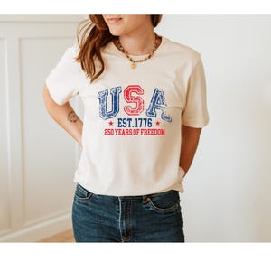 May include: Cream-colored t-shirt with the word "USA" in distressed red and blue lettering. Below the word "USA" is "EST. 1776" and "250 YEARS OF FREEDOM" in red. The shirt is paired with blue jeans.