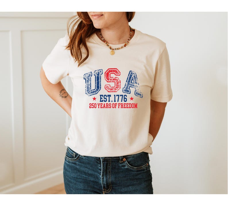 May include: Cream-colored t-shirt with the word "USA" in distressed red and blue lettering. Below the word "USA" is "EST. 1776" and "250 YEARS OF FREEDOM" in red. The shirt is paired with blue jeans.