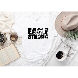 Op de afbeelding: Wit T-shirt met zwarte tekst die "EAGLE STRONG" luidt met een adelaargrafiek in het woord "EAGLE".