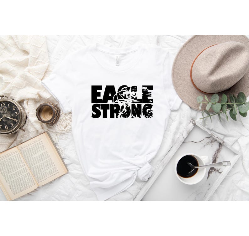 Op de afbeelding: Wit T-shirt met zwarte tekst die "EAGLE STRONG" luidt met een adelaargrafiek in het woord "EAGLE".