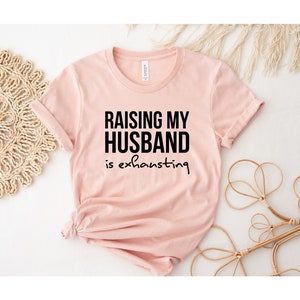 May include: A pink t-shirt with black text that reads "Raising my husband is exhausting".