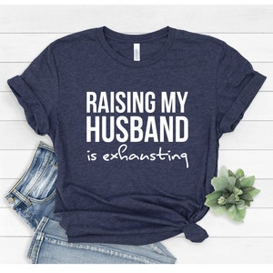 May include: A navy blue t-shirt with the text "Raising my husband is exhausting" in white.