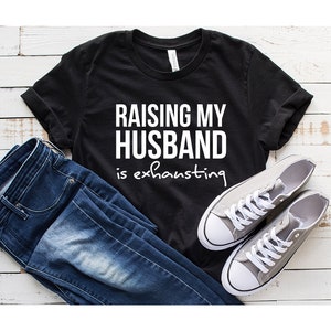 May include: Black t-shirt with white text that reads "Raising my husband is exhausting". The shirt is laid flat on a white wooden surface with a pair of gray sneakers and blue jeans.