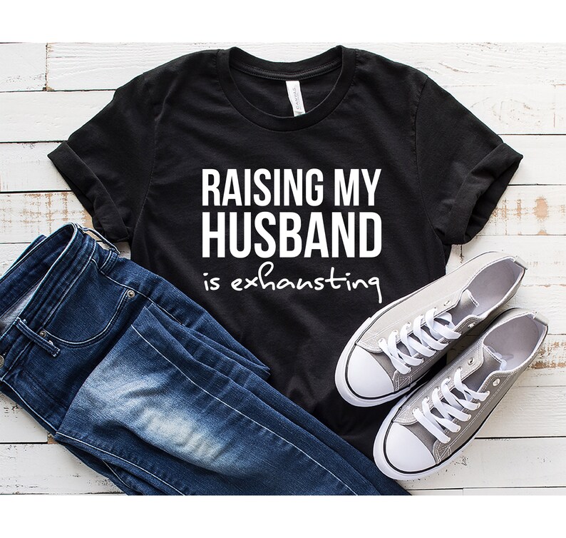 May include: Black t-shirt with white text that reads "Raising my husband is exhausting". The shirt is laid flat on a white wooden surface with a pair of gray sneakers and blue jeans.