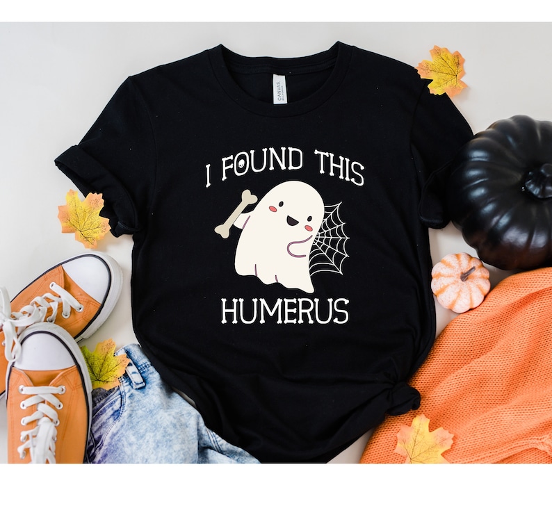 May include: Black t-shirt with a white ghost holding a bone and a spider web. The text on the shirt reads "I found this humerus".