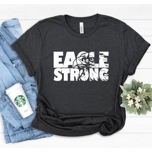 Op de afbeelding: Een donkergrijs T-shirt met een wit grafisch ontwerp dat "EAGLE STRONG" zegt met een adelaarskop in de "O".