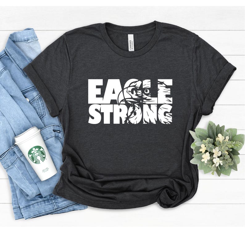 Op de afbeelding: Een donkergrijs T-shirt met een wit grafisch ontwerp dat "EAGLE STRONG" zegt met een adelaarskop in de "O".