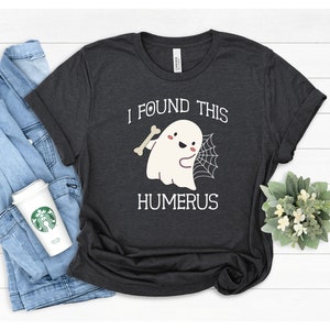 May include: A dark gray t-shirt with a white graphic of a ghost holding a bone and a spider web. The text "I found this humerus" is printed below the ghost.