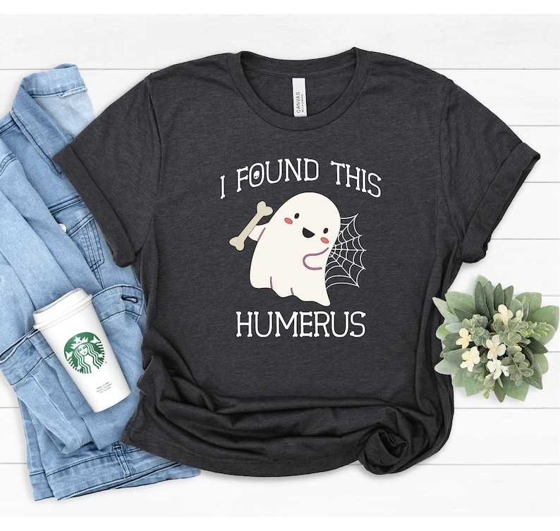May include: A dark gray t-shirt with a white graphic of a ghost holding a bone and a spider web. The text "I found this humerus" is printed below the ghost.