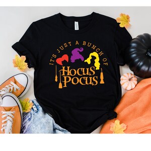 Puede incluir: Camiseta negra con el texto "It's just a bunch of Hocus Pocus" en naranja y amarillo. Tres siluetas de brujas est&aacute;n por encima del texto.
