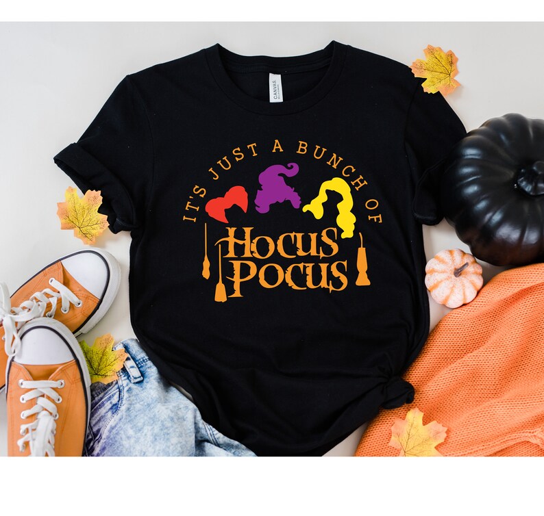 Puede incluir: Camiseta negra con el texto "It's just a bunch of Hocus Pocus" en naranja y amarillo. Tres siluetas de brujas est&aacute;n por encima del texto.