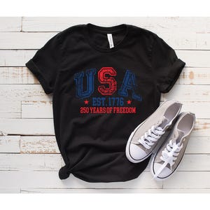 May include: Black t-shirt with the word "USA" in red and blue distressed lettering, along with "EST. 1776" and "250 YEARS OF FREEDOM." Gray canvas sneakers with white laces are next to the shirt.