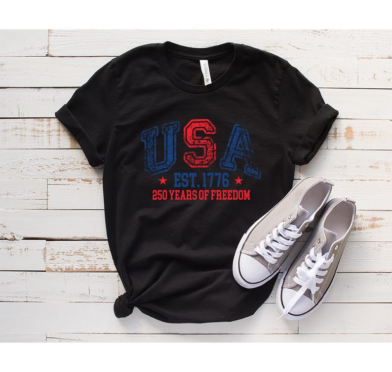 May include: Black t-shirt with the word "USA" in red and blue distressed lettering, along with "EST. 1776" and "250 YEARS OF FREEDOM." Gray canvas sneakers with white laces are next to the shirt.
