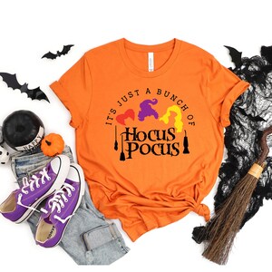 Puede incluir: Camiseta naranja con el texto "It's just a bunch of Hocus Pocus" en letras negras. El texto est&aacute; rodeado por tres siluetas de brujas con cabello morado, amarillo y rojo.
