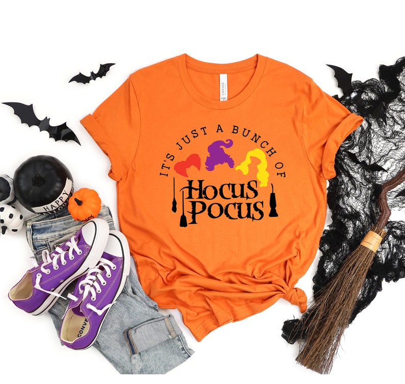 Puede incluir: Camiseta naranja con el texto "It's just a bunch of Hocus Pocus" en letras negras. El texto est&aacute; rodeado por tres siluetas de brujas con cabello morado, amarillo y rojo.