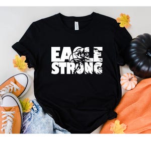Op de afbeelding: Zwart T-shirt met witte tekst die "Eagle Strong" luidt met een adelaargrafiek in het woord "Eagle".