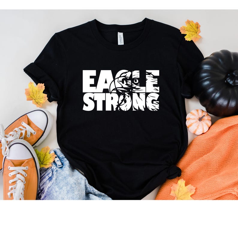 Op de afbeelding: Zwart T-shirt met witte tekst die "Eagle Strong" luidt met een adelaargrafiek in het woord "Eagle".