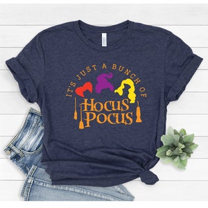 Puede incluir: Una camiseta azul marino con el texto "It's just a bunch of Hocus Pocus" en naranja. Tres siluetas de brujas aparecen en rojo, morado y amarillo.