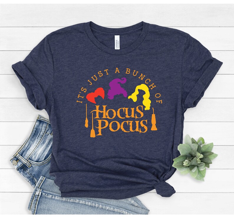 Puede incluir: Una camiseta azul marino con el texto "It's just a bunch of Hocus Pocus" en naranja. Tres siluetas de brujas aparecen en rojo, morado y amarillo.