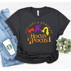 Puede incluir: Una camiseta gris oscuro con el texto "It's just a bunch of Hocus Pocus" en naranja y amarillo. Tres siluetas de brujas est&aacute;n en el centro del texto, con una escoba a cada lado.