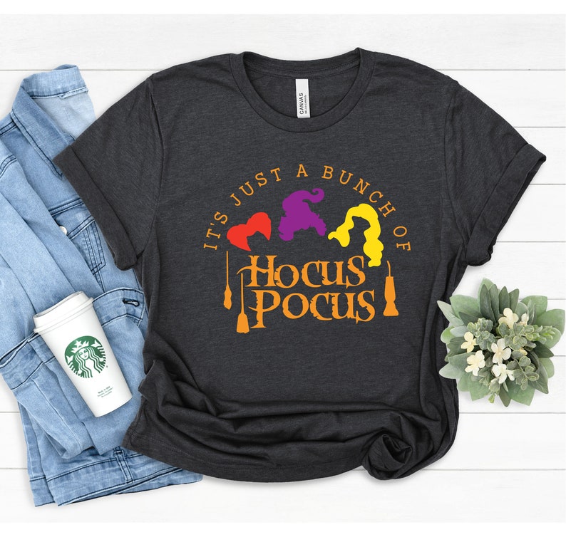 Puede incluir: Una camiseta gris oscuro con el texto "It's just a bunch of Hocus Pocus" en naranja y amarillo. Tres siluetas de brujas est&aacute;n en el centro del texto, con una escoba a cada lado.