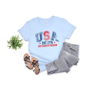 May include: Light blue t-shirt with the word "USA" in red and blue distressed lettering, with "EST. 1776" and "250 YEARS OF FREEDOM" printed below. The shirt is paired with gray jeans and brown sandals.
