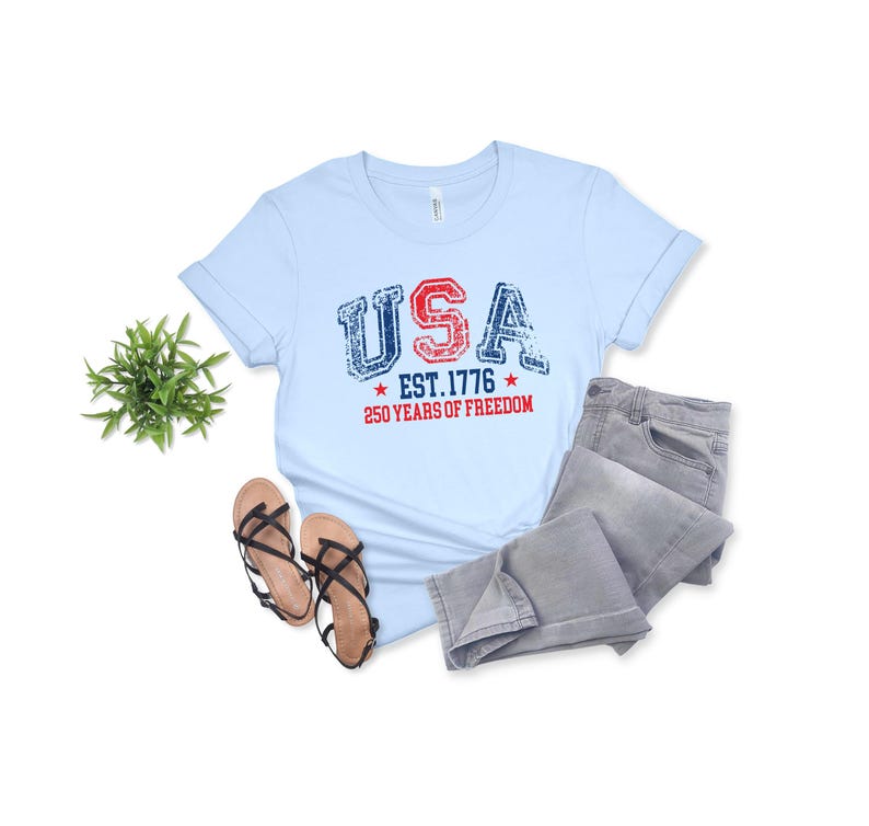 May include: Light blue t-shirt with the word "USA" in red and blue distressed lettering, with "EST. 1776" and "250 YEARS OF FREEDOM" printed below. The shirt is paired with gray jeans and brown sandals.