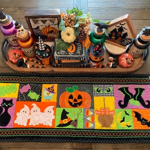 May include: A rectangular Halloween-themed fabric table runner featuring colourful patchwork squares with embroidered designs of a black cat, ghosts, a pumpkin, a witch hat, a candy apple, and a bat, displayed with a wooden tray of seasonal decor.