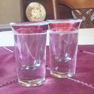 Puede incluir: Dos vasos de chupito transparentes con borde rosa y banda blanca. Los vasos tienen fondo redondeado y están llenos de líquido transparente. Los vasos están sobre una superficie morada.