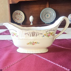May include: Cream-colored gravy boat with a decorative floral pattern in pink, yellow, and blue. The boat has a curved handle and a gold trim. Other decorative plates and an egg-shaped ornament are in the background.