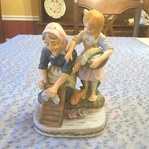 Puede incluir: Una figura de cerámica que representa a una anciana lavando ropa con una niña que sostiene una cesta. Las figuras visten ropa vintage, con la anciana con un pañuelo en la cabeza y un delantal. La base presenta un diseño de agua y flores.