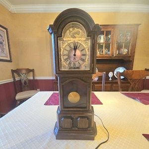 Puede incluir: Un reloj de pie alto de madera oscura con una esfera de color claro y números romanos. El reloj tiene un acabado marrón oscuro y una parte superior decorativa. El reloj está sobre un mantel blanco.