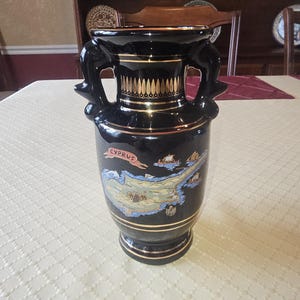May include: A black ceramic vase with gold accents and two decorative handles. The vase features a map of Cyprus and the word "CYPRUS" in a banner. The vase is on a white tablecloth.