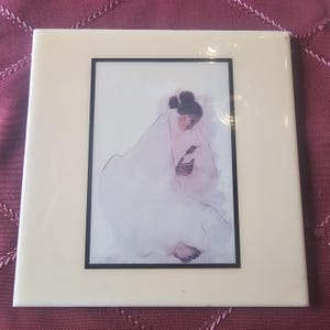 May include: A framed art print with a watercolour illustration of a person in white fabric. The artwork is within a black border and a cream frame. The image is on a burgundy surface.