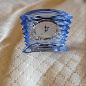 Könnte beinhalten: Eine blaue Glasuhr mit silbernem Zifferblatt. Die Uhr hat ein einzigartiges, geriffeltes Design und steht auf einer cremefarbenen Oberfläche. Das Zifferblatt hat schwarze Zeiger und kleine schwarze Punkte für die Zahlen. Maße in Zentimetern.